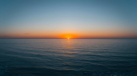 The sun is setting on the horizon, casting a warm orange glow across the sky and reflecting on the calm, deep blue ocean. The water is mostly still with a few gentle ripples. The sky transitions from a pale blue at the top to a soft orange near the horizon. This image evokes a sense of peace and vastness.の写真素材
