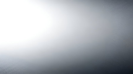 An abstract background featuring a smooth silver gradient that transitions from bright white on the left to a darker gray on the right. Subtle, fine speckles are visible across the surface, adding a delicate texture to the otherwise smooth appearance.の写真素材