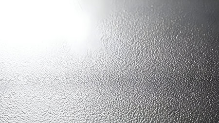 A detailed macro shot of a textured metallic surface. The texture is granular with fine crack-like lines visible across the surface. A bright light source reflects from the upper left corner, casting a gradient of light and shadow across the metallic material, creating a subtle and abstract background.の写真素材