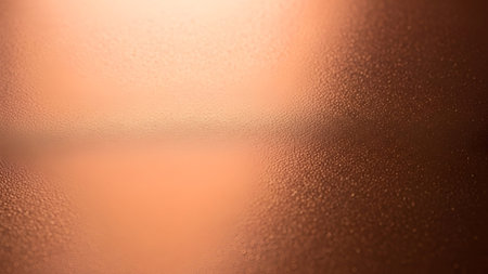 A close-up, abstract view of a frosted copper surface covered in tiny water droplets. The texture is detailed and granular, with a warm, gradient of orange and brown tones. Light reflects off the surface, highlighting the condensation and creating a sense of depth and moisture.の写真素材