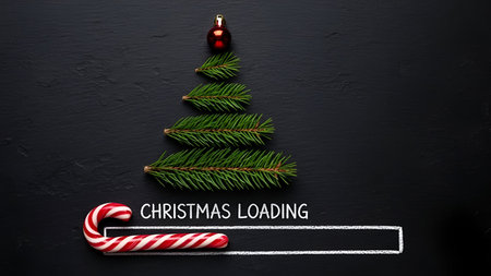 The a minimalist Christmas tree constructed from three horizontal pine branches arranged in a triangular shape. At the of the tree, there is a small red Christmas bauble. Below the tree, there is a candy cane with a white loading bar that has the text "CHRISTMAS LOADING" written in white. The entire composition is set against a dark, textured black background. The conveys a festive and holiday mood with a focus on simplicity and minimalism.の写真素材