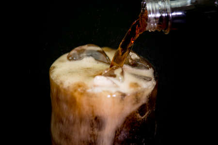 Pouring cola from plastic bottle into rocks glass with ice cubes at black backgroundの写真素材