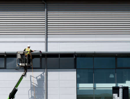Construction worker on mobile boom lift cleaning external rainscreen cladding on new build office and warehouse developmentのeditorial素材