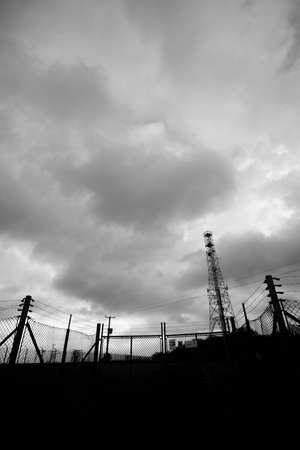 Monochrome silhouetted telecommunications mast with security fencing and gate against a cloudy skyの写真素材