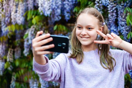Portrait of cute preteen caucasian girl with ponytails taking selfie against blooming wisteria.の写真素材