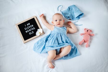 Eleven months old baby girl wearing blue summer dress laying down on white background with letter...