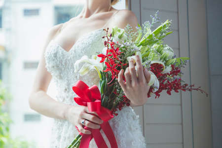 Women blonde hair wear wedding blue rings in white wedding dress. Holds a red and white bouquet and red ribbon by the window. White background and bright style.の写真素材