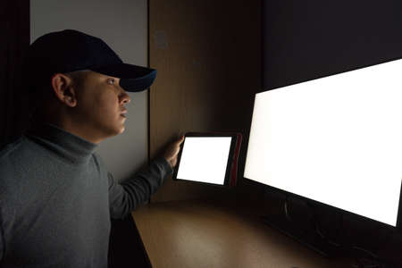 Side view of man Hacker sit at the computer monitor, white screen tablet in the dark room.