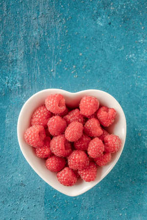 ripe red raspberry fruits in a heart shaped bowl on a blue backgroundの写真素材