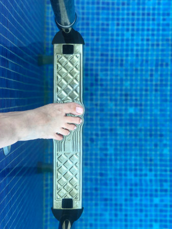 pool, closeup top view female foot steps into blue swimming pool with ladder in summer. summer sport or recreation activity in a resort or spa center. summer holiday concept with woman body partの写真素材