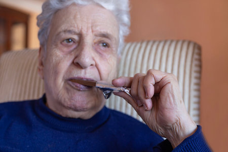 selective focused wrinkled hand of woman eating delicious chocolate bar. old woman or man eating junk food. hand holds chocolate bar to eat. senior person biting sweet foodの写真素材