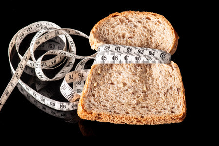 slice of bread tied with measurement tape on black. Diet concept background or surface. piece of wholegrain bread grasped by measuring tape isolated on black. healthy eating habit. healthy food ideaの写真素材