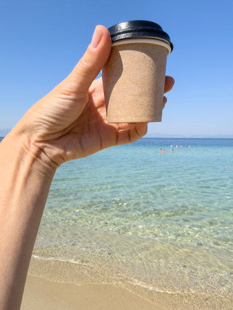 take away coffee, female hand holds takeaway coffee or tea cup over beautiful sea under blue sky with copy space in sunny summer day. hot drink or beverage background in summer. blank paper mug or cupの写真素材