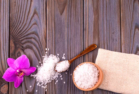 White bath salt in a wooden bowl with a spoon, soap, burlap and orchid on wooden tableの写真素材