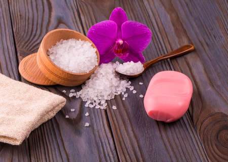 White bath salt in a wooden bowl with a spoon, soap, burlap and orchid on wooden tableの写真素材
