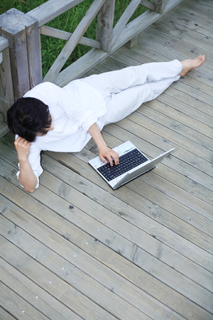 Young Asian man lying on the wooden bridge, working on his laptopの写真素材