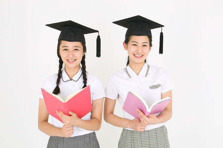Two Asian girl students wearing a graduation hat and and reading a book in the studio, isolated on whiteの写真素材