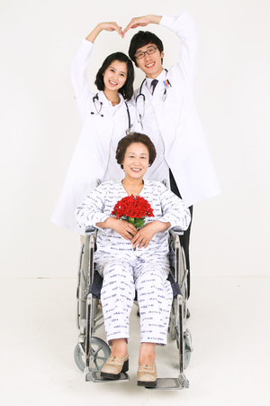 A couple of doctors making heart with arms as standing behind the senior patient in wheelchairの写真素材