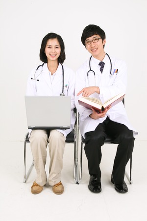 Full body shot of a female doctor working on a laptop as sitting next to a male doctor who is holding a bookの写真素材