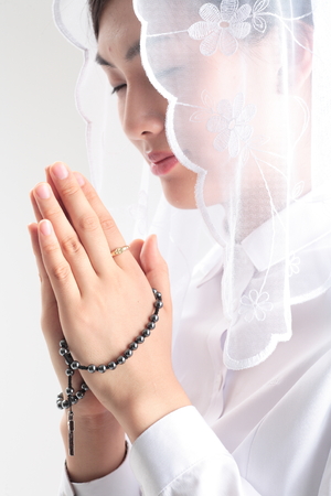 Close up shot of A female Asian wearing a mass veil praying with toothy smile as holding a rosary in handの写真素材
