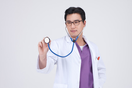 Asian male doctor wearing robe isolated in white - pretending to apply stehoscope in the airの写真素材