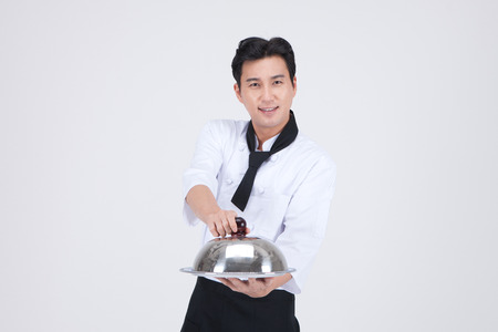 Asian cook wearing black apron isolated in white - holding a plate with dome coverの写真素材
