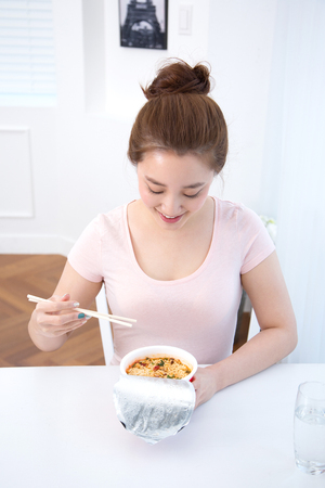 Young female Asian eating noodle cup/cup noodles in white interior roomの写真素材