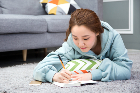 Young female Asian in pyjamas writing down as lying down on living room floorの写真素材