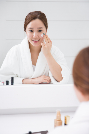 Young female Asian in white shower robe applying skin care lotion/cream in bathroomの写真素材