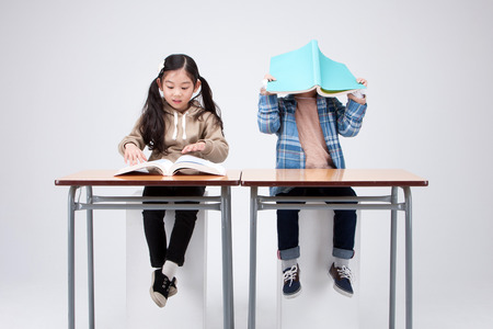 Isolated shot in studio - little Asian girl and boy sitting at desk and posing togetherの写真素材