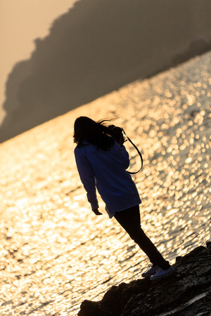 Back shot of a woman holding a camera on the rocky beach at sunsetの写真素材