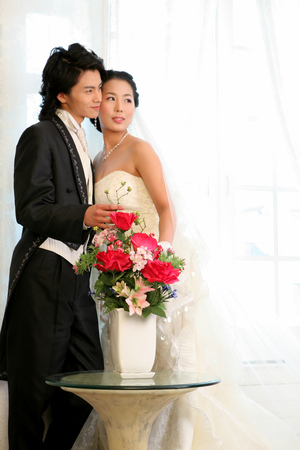 Asian bride and groom in wedding dresses posing in a studio with flowers in vaseの写真素材