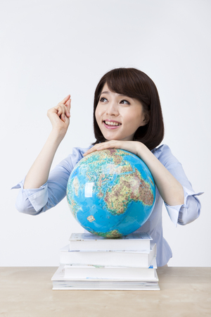 Portrait of Asian female teacher leaning on globe and pile of books on the desk isolated on whiteの写真素材