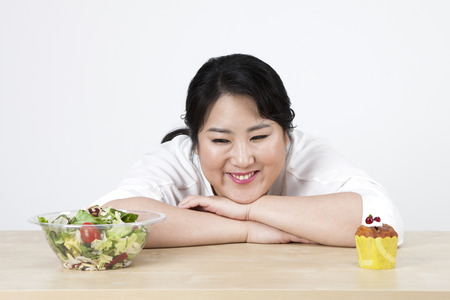 Asian overweight woman choosing whether salad or cupcake isolated on whiteの写真素材
