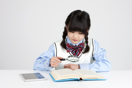 Asian elementary school girl looking through book with magnifier isolated on whiteの写真素材
