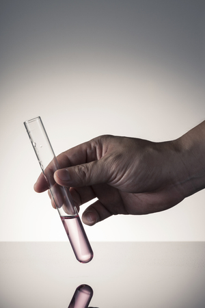 A test tube filled with red solution on grey background is held by a right hand.の写真素材