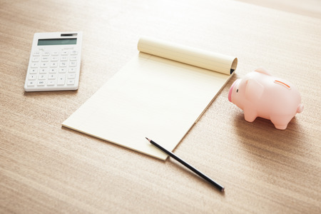 A piggy bank, a notepad, a pencil, and a calculator on brown backgroundの写真素材