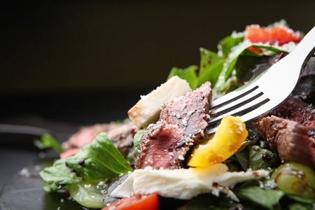 Close-up of a fork with beef salad which has sprinkled cheese on a black plateの写真素材