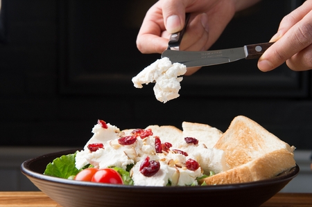 A close-up shot of hands holding a knife and a fork, making a ricotta cheese salad with bread and cranberryの写真素材