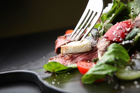 Fork with a beef salad with tomatoes, green grapes and sprinkled cheese on a black plate, close-upの写真素材