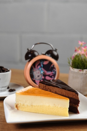 Piece of cheesecake and a piece of chocolate moose cake on white plate, on wooden table, with an alarm clockの写真素材