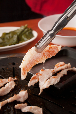 Tongs turning pork cheek meat, being grilled, on a black grillの写真素材