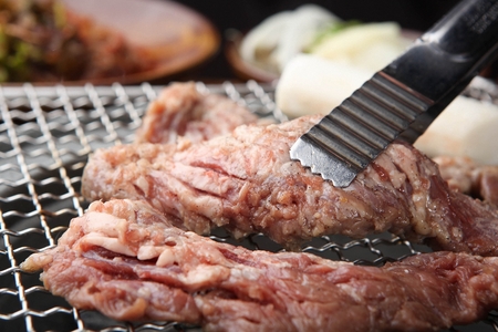 Tongs grabbing pork skirtmeat, being grilled, on a grill placed over charcoalの写真素材
