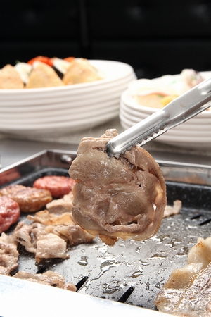 Tongs grabbing pork neck slice, being grilled, on a black grillの写真素材
