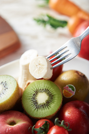 Fork lifting a banana from a platter of kiwis, apples, bananas, cherry tomatoesの写真素材