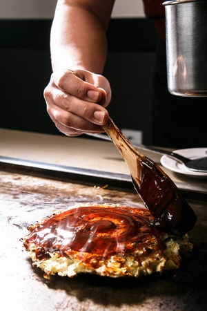 Chef putting teriyaki sauce on Okonomiyaki, japanese pancakes with dried bonito, mayonnaise, teriyaki sauce, on iron plateの写真素材