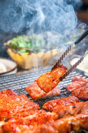 Pincers picking up red chili pepper paste-marinated pork belly being grilled on charcoal grateの写真素材