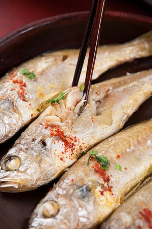 Chopsticks picking up grilled yellow croaker with sprinkled chili powder and chives, on round plate