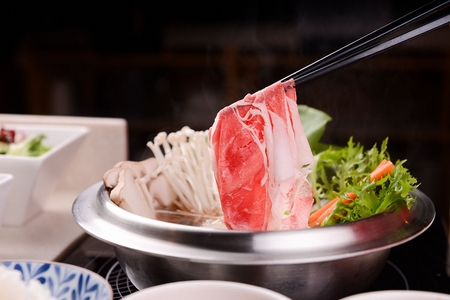 Chopsticks picking up meat from shabu-shabu made of various vegetables such as mushrooms and pak choi, with glass noodles, in pot