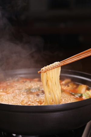 Chopsticks grabbing the noodles from spicy chopped noodles soup with mushrooms and crown daisy, in potの写真素材